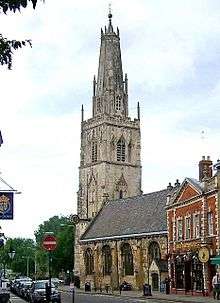 A stone church in a town seen from the southeast. St the far end is a large tower surmounted by a truncated spire with pinnacles and, at the top, a ball finial