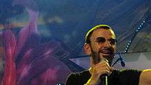 A colour photograph of Starr wearing sunglasses and singing into a microphone. The background is blue and purple.