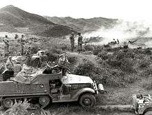 A half track vehicle sits in the foreground in a grassy open area, with a large calibre machine-gun mounted on top. Soldiers using binoculars are sitting on the vehicle, while in the background smoke obscures a number of artillery guns which are firing