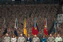 A stadium full of soldiers sits behind a podium of commanders in military uniform