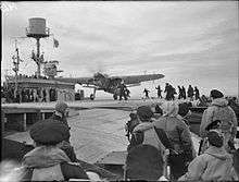 Black and white photograph of a single-engined monoplane on the flight deck of a World War II-era aircraft carrier. Large numbers of men are watching the aircraft or running towards it.