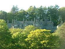 Peeping over the tops of trees in the foreground is a stone house with five gables of different sizes. More trees are in the background.