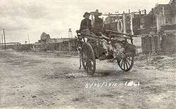Two men in sombreros riding in a donkey-cart with a line of feet sticking out the back. They are riding down a dirt street away from the camera, with a line of buildings on the right. Dated 5/15/1911.