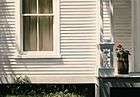 oil painting, close-up;old clapboard white house with partial golden curtains in window on left, porch on right with geranium plant in wooden barrel