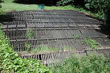 A horizontal wooden rack roughly 30 feet (9.1 m) wide and 30 feet (9.1 m) long lies in flat, grassy area.