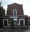 A plain building in very dark brick. In the foreground is a flat-roofed entrance area with two wooden doors and a small window (partly obscured). Behind this, a squat tower, with a central window, rises in several stages.