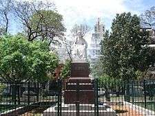 A statue in a plaza, with trees at the sides, and wires around it.