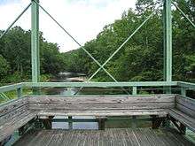 A detail of the wooden seat and observation platform at the center of the green Whipple truss bridge facing out over a river