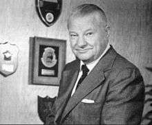 Black-and-white portrait photograph of gray-haired man in suit posing to the right; a number of plaques are present on the wall behind.