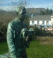 In the foreground is a dark-coloured statue of a man carrying a small dog. In the background is a low, white building with cars parked outside.