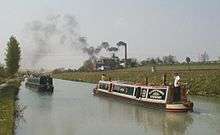 A straight section of canal going into the distance, with two passing narrow boats in the foreground, with a large brick building and tall chimney in the middle-distance on the right bank of the canal. Smoke is billowing from the chimney, blowing across the canal to the left.