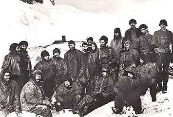 A group of men sitting closely packed together, in heavy winter clothes and wearing hats. Snow and ice on the ground and in the background