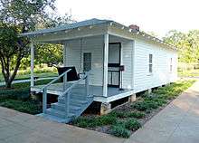 Present-day photograph of a whitewashed house, about 15 feet wide. Four bannistered steps in the foreground lead up to a roofed porch that holds a swing wide enough for two. The front of the house has a door and a single-paned window. The visible side of the house, about 30 feet long, has double-paned windows.