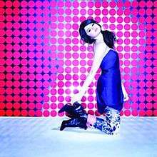 A young woman wearing a blue shirt kneels and poses during a photo-shoot, wearing zebra-printed pants, and high-top boots. The floor is metallic, and the background is filled with several small pink circles.