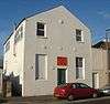 Three-quarter view of a tall, two-storey, plain-walled, white-painted building with a pointed roof. The front has one blank window and a half-blank, half-glazed window flanking a narrow door, above which is an orange sign. There are also six circular protrusions. The side wall has three windows and a thin metal flue.