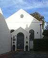 A white stuccoed hall, partly in shadow and linked at its left corner to the wall of a larger building, which partly obscures it. An entrance porch has a grey double-door and a semicircular transom light, two lamps and a plaque reading "Gospel Hall". The porch obscures two semicircular-arched windows, above which is a pediment with a round window.