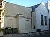 Side view of a two-part, plain-walled building in shadow. The nearer section is flat-roofed, painted cream and has two blank windows with a wide pillar between them.  Rubbish bins and scaffolding are to the left.  The far section is white, has a pointed roof whose end resembles a pediment, and has two identical red-framed windows.