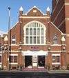 Front view of a squat, red-brick building with a steep pointed roof and prominent stone dressings. There are four identical spirelets: two ending about halfway up the building and two level with the top of the roof. A pointed-arched window, split by mullions into ten panes, dominates the façade. Below it are two entrance doors and the word "ten" in purple lower-case letters.
