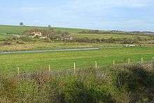 Photograph of grassland bisected by a canal, with farm buildings in the distances and the bushes of a railway cutting in the foreground.