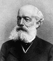 A black and white image of a bald man in a dark outfit, with a bushy white beard and moustache
