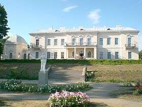 Large, ornate building with large windows, a columned portico, and iron filigree balconies, sits on a hill.