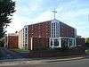 A shallow-roofed building with an extension at the left rear. The façade has small, regularly spaced concrete protrusions, but is mostly brick. In the centre of this section is a tall 4x5 window extending to the roofline. A wide, flat-roofed entrance area sits in front of this. A series of angled ground-to-roof windows run down both sides.