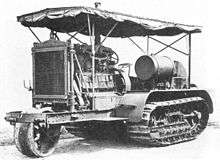 An early bulldozer-like tractor, on crawler tracks, with a leading single wheel for steering - projecting from the front - on an extension to the frame. The large internal combustion engine is in full view, with the cooling radiator prominent at the front. An overall roof is supported by thin rods, and side protection sheeting is rolled up under the edge of the roof.