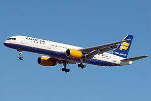 A mostly white Boeing 757 with blue and yellow trim preparing for landing against a blue sky. Landing gear and flaps are fully extended in final approach configuration.