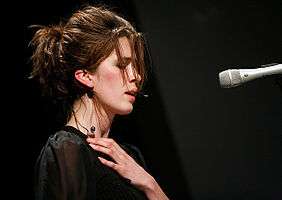 Imogen Heap face-on, on 12 October 2006 at the Carling Academy Birmingham, at a multi-tier set-up with keyboard atop which she's intently playing. She has a headset microphone, then-characteristically back-combed hair, wears a sleeveless frilly pink bodice and is rear illuminated by two piercing green star-burst spotlights.