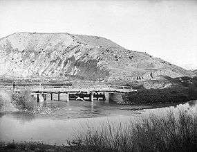 A photo of a river in the foreground and hills in the background. A small dam or diversion is in the center of the photo where a canal flows off from the river.