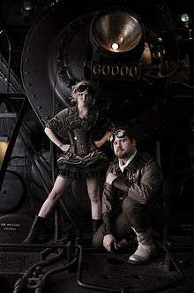 A short skirted women stands assertively behind a sitting man in front of a large machine
