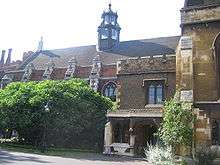 A long, low building of brick and stonework, with arched windows. It is partially obscured by trees and shrubs. The slate roof features a six-sided tower of stone and class.