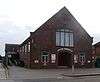 Three-quarter view of a dark brick building with lighter brick dressings around the arched entrance. "LANCING TABERNACLE" is written in white letters above this and below a wide, tall, slightly arched window with brown mullions. Evenly-spaced rectangular windows extend down the longer side. A brick-paved area is in front.