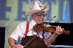 An elderlyl Luderin Darbone playing the fiddle. He is sitting and wearing a cowboy hat.