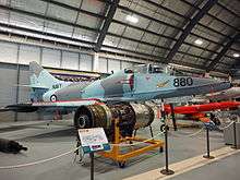 Colour photograph of a military fighter jet aircraft painted in blue and grey camouflage in a museum other aircraft and engines are visible.