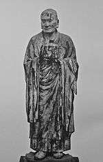 Muchaku (Asanga). Frontal view of a lifelike statue keeping his hands in front of his body with one palm turned up and the other facing sidewards as if keeping something. Black and white photograph.