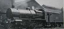 Side-and-front view of a modified version of the N class on shed. The distinguishing feature from normal N class locomotives is the tall cylindrical chimney on the smokebox. A member of the crew is standing next to the tender.