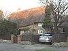 A wide stone cottage with a deep thatched roof illuminated by low sunlight. Two small rectangular windows can be seen on the right of the building, next to some trees and a car. A bush, fence and the roof obscure most of the rest of the cottage. There is a small chimney-stack on the left.