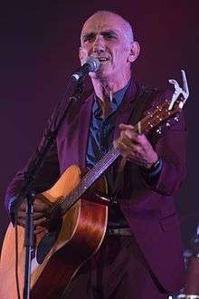 Head and shoulders of a fifty-two-year-old male standing at a microphone. He is shown in right profile, wearing a harmonica in a cradle and staring into the distance.