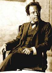 Middle-aged man, seated, facing towards the left with head turned towards the right. He has a high forehead and rimless glasses, and is wearing a dark, crumpled suit