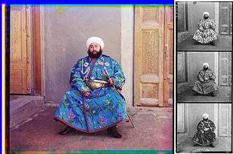 A large color photograph abutting (to its right) a column of three stacked black-and-white versions of the same picture. Each of the three smaller black-and-white photos are slightly different, due to the effect of the color filter used. Each of the four photographs differ only in color and depict a turbaned and bearded man, sitting in the corner an empty room, with an open door to his right and a closed door to his left. The man is wearing an ornate full-length blue robe trimmed with a checkered red-and-black ribbon. The blue fabric is festooned with depictions of stems of white, purple, and blue flowers. He wears an ornate gold belt, and in his left hand he holds a gold sword and scabbard. Under his right shoulder strap is a white aiguillette; attached to his robe across his upper chest are four multi-pointed badges of various shapes, perhaps military or royal decorations.