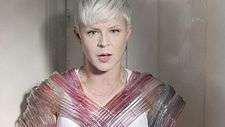 A woman with platinum blonde hair is standing alone in front of a wall, wearing a dress with plastic tubes, filled with red and blue liquid, wrapped around her body.