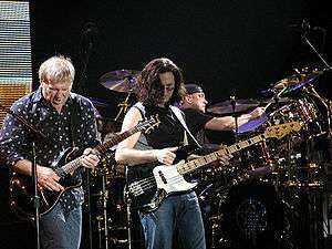 Color photo of 3 musicians on a stage, in the foreground, one man is holding a guitar, while the other is holding a bass guitar, and in the background a man playing drums.