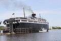 A large black ship with white upper decks rests at a berth, its rear surrounded by wooden posts. The rear access to its vehicle decks is opened upward.