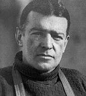 Head and shoulders of a dark-haired man looking directly to camers. The straps of a harness over his shoulders are visible.