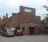 A brick building with an angular façade consisting of stepped brick walls above an entrance porch with a wooden door in a recess. A crucifix and the words "SHOREHAM FREE CHURCH" are on the façade.