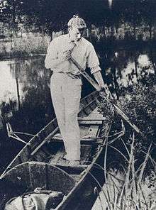 Paget standing in a canoe wearing a deerstalker cap, holding an oar