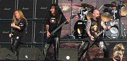 Three men on a stage, all holding guitars. All three are wearing black clothing, and audio equipment can be seen both in front of and behind them.