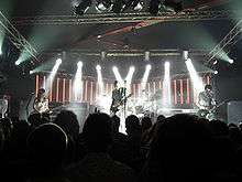 The Smashing Pumpkins perform on a back-lit stage dressed in white. From left to right: Ginger Pooley—a brunette Hispanic woman in her 20s wearing a sequined dress—looks at her bass guitar, Billy Corgan—a bald, middle-aged Caucasian man with a white jumpsuit and a black-and-white striped T-shirt—plays guitar, Jimmy Chamberlin plays a silver drum kit obscured behind Corgan, and Jeff Schroeder—a Korean male in his 30s with mid-length brunette hair—looks at his guitar while playing.