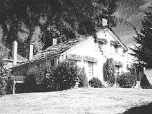 A modest two-story farmhouse with a peaked roof and four chimneys sits under a large tree at the top of a sloping lawn. Bushes and shrubs grow near the house on its two visible sides. A small part of a neighboring house, perhaps only 15 feet (4.6 m) away, can be seen in the background.